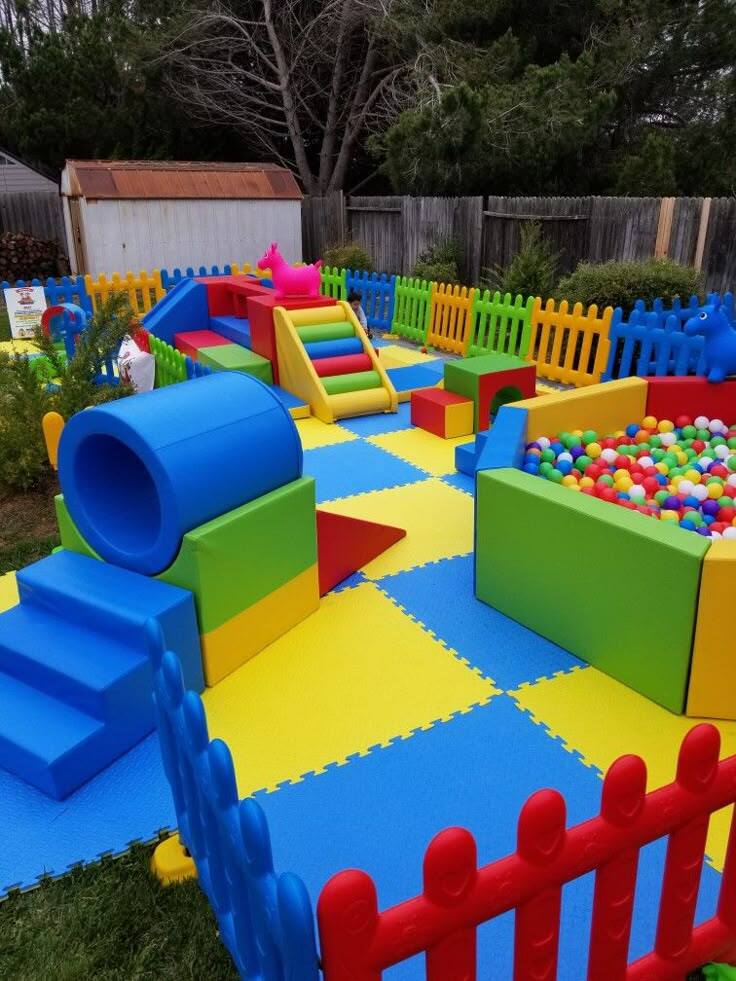 Colorful soft play birthday party setup outdoors with foam blocks, slides, ball pit and fence enclosure for toddlers