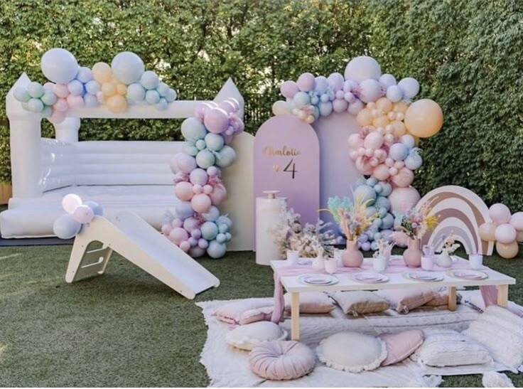 Driveway first birthday party setup with pastel balloon arch, white bounce house and boho picnic table
