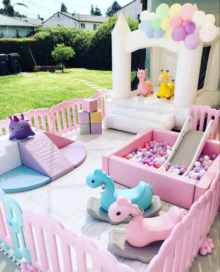 Pastel backyard toddler birthday party setup with white bounce castle, pink ball pit, slide and animal toys on grass in Irvine